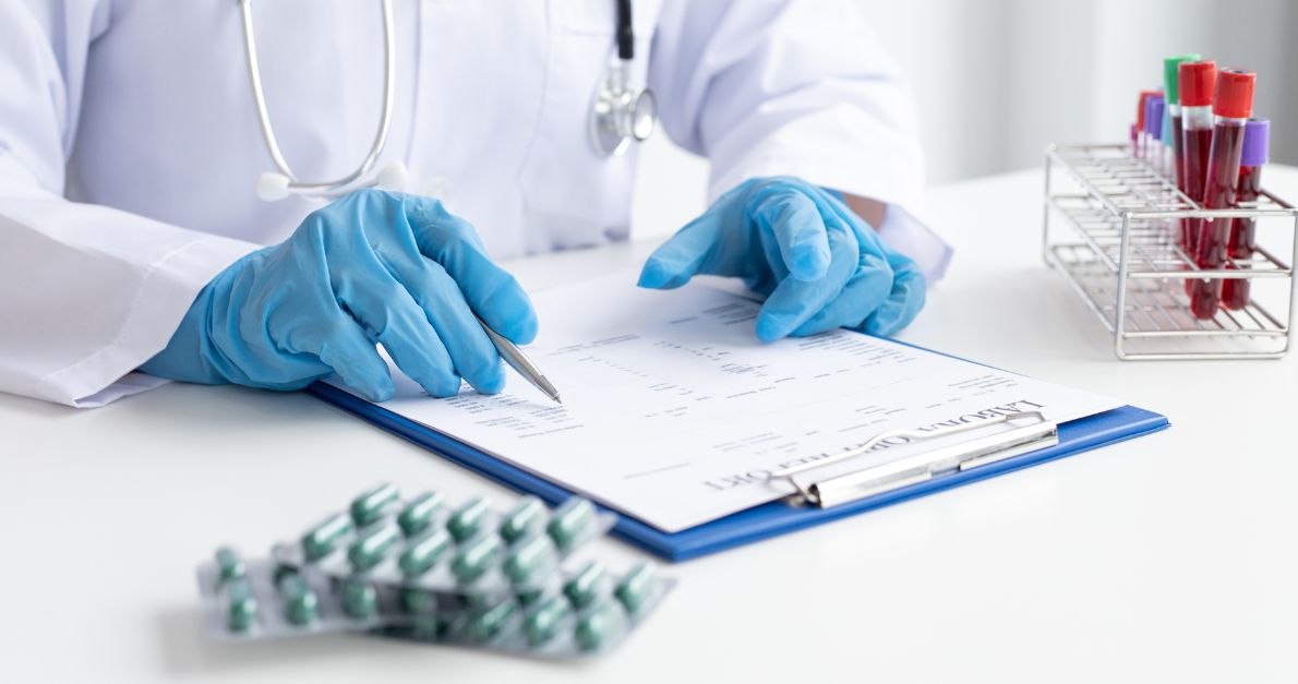 Clinician in blue gloves completing a patient report form on a clipboard, with test tubes in a rack and a blister pack of tablets on the desk.